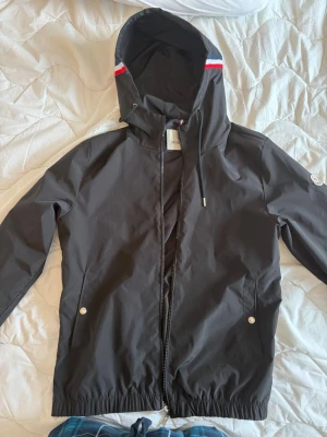 Svart vindjacka från Moncler - Snygg svart vindjacka från Moncler med huva och dragkedja framtill. Jackan har Moncler-logga på ärmen och detaljer i rött, vitt och blått på huvan. Två fickor med tryckknappar och justerbar dragsko i huvan. Perfekt för blåsiga dagar. Passar S-M