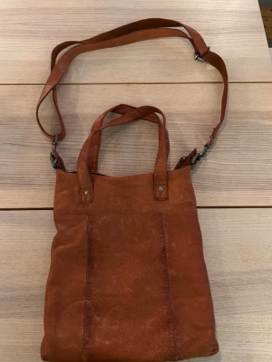 Brun axelväska i skinn - Snygg brun axelväska i skinn med justerbar och avtagbar axelrem och två korta handtag. Väskan har en enkel, stilren design och passar perfekt för dig som vill ha en praktisk och trendig väska till vardagen. Perfekt storlek för det viktigaste och lätt att bära med sig. Väskan har ett fack med dragkedja, i vilket det finns ytterligare små fack varav det ena har dragkedja.  Mycket gott skick.  Mått: 30x33 cm