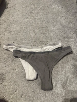 Två SKIMS stringtrosor grå & vit - Säljer två snygga stringtrosor från SKIMS i färgerna grå och vit. Båda har en elastisk midjeresår med SKIMS-logga och är tillverkade i mjukt, stretchigt material som sitter skönt på kroppen. Perfekta för dig som gillar minimalistisk och stilren design.