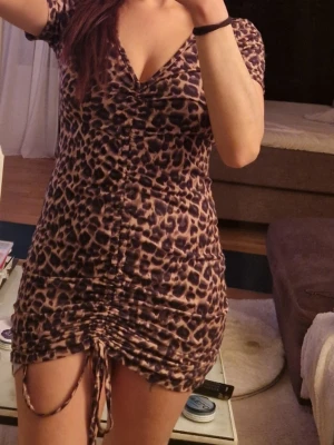 Leopardmönstrad kort klänning - Säljer en kort, tight klänning med leopardmönster och snörning framtill. Klänningen har v-ringning och korta ärmar. Perfekt för dig som vill sticka ut med ett djuriskt mönster. Materialet känns mjukt och följsamt.