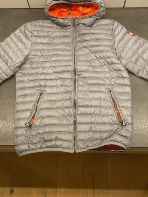 Ljusgrå Colmar pufferjacka 14Y - Säljer en ljusgrå pufferjacka från Colmar med orange foder och huva. Jackan har två fickor med dragkedja och en snygg quiltad design. Perfekt för dig som vill ha en lätt och stilren jacka med sportig känsla.