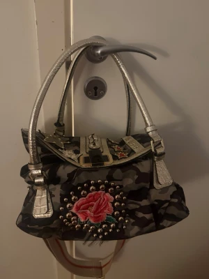 Replay handväska med camouflage och stjärnor - Unik handväska från Replay med camouflage-mönster, silverfärgade detaljer och handtag. Väskan har broderad röd ros, nitar och flera stjärnpatches i olika färger. Axelrem i tyg och coolt spänne framtill. Perfekt för dig som vill sticka ut! Köptes för 2500 kr säljer för 1000