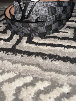 Rutig  bälte i svart - Säljer ett svart och grått bälte  med klassiskt rutigt mönster och stor-logga i metall framtill. Bältet har en stilren design och är gjort i ett tåligt material som känns lyxigt. Perfekt accessoar för att lyfta vilken outfit som helst.