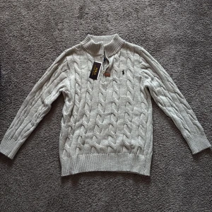 Grå stickad tröja från Polo Ralph Lauren - Säljer en stilren grå stickad tröja från Polo Ralph Lauren med kabelstickat mönster och dragkedja vid halsen. Tröjan har långa ärmar och ribbade muddar. Perfekt för kyliga dagar