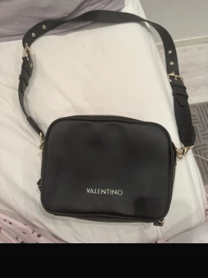 Svart axelväska från Valentino - Snygg svart axelväska från Valentino med guldfärgade detaljer och justerbar axelrem. Väskan har en stilren, fyrkantig form och dragkedja upptill. Insidan har ett fack med dragkedja och logotyp. Perfekt för att bära det viktigaste.