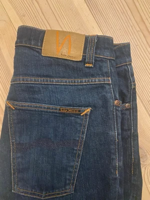  Nudie Jeans Co. Mörkblå - Snygga mörkblå jeans från Nudie Jeans Co modell thin finn med klassisk femficksdesign och orange kontrastsömmar. Modellen har rak passform, knappgylf och läderpatch med logga bak. Perfekta för dig som gillar stilrena och tidlösa jeans. Fråga på bara!