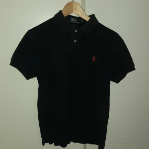 Svart piké från Polo Ralph Lauren - Klassisk svart pikétröja från Polo Ralph Lauren med röd broderad logga på bröstet. Tröjan har korta ärmar, krage och knappar framtill. Tillverkad i mjuk bomull för en skön känsla och snygg passform.