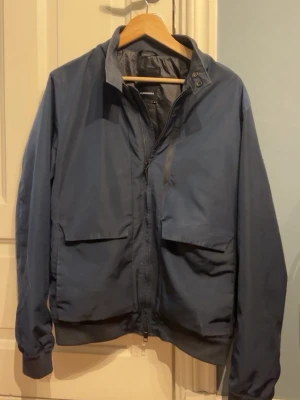  J.Lindeberg Jacka  - Säljer en stilren mörkblå vindjacka från J.Lindeberg. Jackan har dragkedja framtill, stora fickor med lock och en diskret logga på ärmen. Perfekt för blåsiga dagar och enkel att matcha med olika outfits. Klassisk krage och ribbade muddar vid ärmslut.