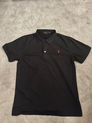 Svart piké från Polo Ralph Lauren - Klassisk svart pikétröja från Polo Ralph Lauren med röd broderad logga på bröstet. Tröjan har krage, två knappar framtill och korta ärmar. Tillverkad i bomull för en skön och luftig känsla.