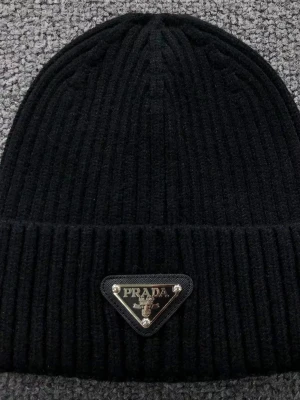   Svart ribbad mössa från Prada  - Svart mössa från Prada med ribbad struktur och klassisk uppvikt kant. Mössan har en metallplakett med Prada-logga framtill och är tillverkad i mjukt material som känns lyxigt och värmande. Perfekt accessoar för att lyfta din vinterstil.
