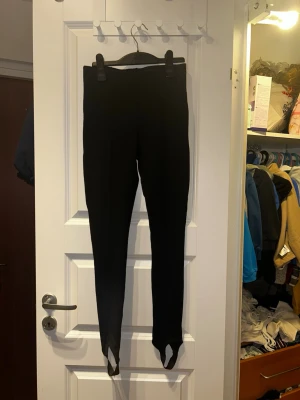 Svarta ridinspirerade leggings Gina Tricot XS - Svarta leggings från Gina Tricot i storlek XS med klassisk ridbyx-look och fotstrappar. Hög midja och tight passform, sydda pressveck framtill. Materialmix av viskos, polyester och elastan för stretch och komfort.