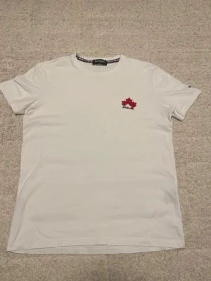 Vit DSQUARED2 t-shirt med lönnlöv - Vit t-shirt från DSQUARED2 i bomull med broderat rött lönnlöv och DSQ2-logga på bröstet. Klassisk rund halsringning och korta ärmar. Snygg detalj med DSQ2-tryck även på ärmen. Perfekt för dig som gillar stilrena plagg med en twist.