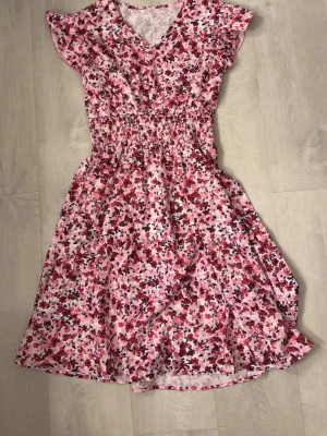 Blommig rosa sommarklänning - Söt blommig klänning i rosa, rött och vitt med v-ringning och korta volangärmar. Smockad midja ger en snygg siluett och kjolen har ett lätt utställd fall. Perfekt för soliga dagar och picknickar.