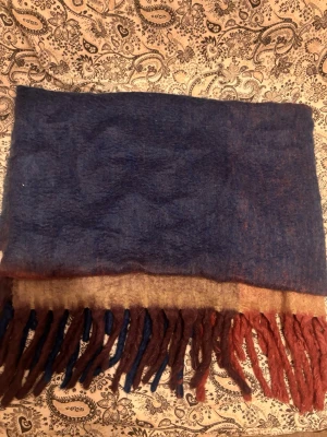 Flerfärgad pläd från Rusta Home - Mysig pläd i blockfärger med blått, beige och vinrött. Pläden har långa fransar nedtill och är tillverkad i en mjuk blandning av akryl, polyester och ull. 