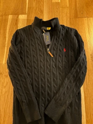 Svart stickad tröja Polo Ralph Lauren - Säljer en svart kabelstickad tröja från Polo Ralph Lauren med halv dragkedja och klassisk röd logga på bröstet. Tröjan har hög krage och långa ärmar, perfekt för kyliga dagar. Materialet är mjukt och värmande ull.