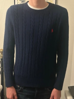 Mörkblå kabelstickad tröja Polo Ralph Lauren - Kabelstickad mörkblå tröja från Polo Ralph Lauren med klassisk röd broderad logga på bröstet. Tröjan har rund halsringning, ribbade muddar och är långärmad. Perfekt för dig som vill ha en stilren och tidlös look.