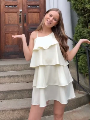 Ljus volangklänning med asymmetrisk axel - Superfin ljus kortklänning med flera volanger och en axelbandslös design på ena sidan. Klänningen har tunna axelband och är tillverkad i ett lätt, flowy material som ger en luftig känsla. Perfekt för sommarens alla tillfällen!