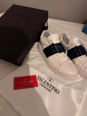 Valentino Garavani vita/mörkblåa - Säljer ett par Valentino Garavani sneakers i storlek 42 👟✨ Skorna är vita med mörkblå detaljer och har endast använts vid ett fåtal tillfällen. De är i mycket gott skick, med välbevarat läder samt sula.  Skorna säljs som ett komplett set 📦 och levereras med originalkartong, dustbag samt kort enligt bilder. Modellen är normal i storleken och passar utmärkt både till vardags och till mer uppklädda tillfällen.  Vid seriöst intresse är du välkommen att höra av dig 💬 Pris kan diskuteras vid bra affär