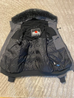 Mörkgrå Zanetti pufferjacka med päls - Säljer en mörkgrå pufferjacka från Zanetti Canada med svart fuskpäls på huvan och flera praktiska fickor. Jackan har ribbade muddar, patch på ärmen och stängs med dragkedja och knappar. Perfekt för kalla dagar och riktigt snygg streetstil ny fick som pressent men vill inte ha den passar inte 