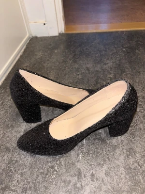 Svarta glittriga pumps med klack - Säljer ett par svarta pumps med glittrig yta och chunky klack. Skorna har spetsig tå och är täckta med glitter som ger en riktigt snygg look. Perfekta för dig som vill sticka ut lite extra. Storlek 38 men passar 39 också