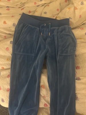 Blå mjukisbyxor i sammet - Säljer ett par blå mjukisbyxor från Juicy Couture i mjuk sammet. Byxorna har bred resår i midjan med snörning, stora fickor fram och bakfickor med knappar. Diskret Juicy Couture-logga broderad på ena fickan framtill. Perfekta för chill dagar.