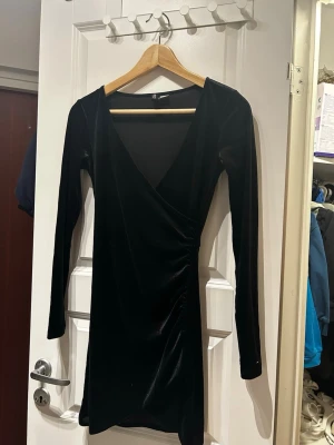 Svart sammetsklänning H&M omlott - Säljer en svart kort sammetsklänning från H&M med omlottdesign och v-ringning. Klänningen har långa ärmar och snygg rynkning i sidan som ger en smickrande siluett. Perfekt för dig som vill ha en stilren och elegant look.