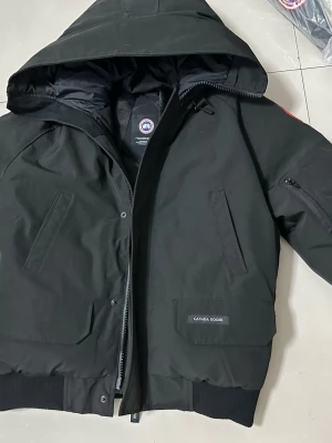 Svart dunjacka från Canada Goose Size S - Säljer en svart dunjacka från Canada Goose med huva och flera fickor. Jackan har dragkedja och tryckknappar framtill, ribbade muddar och klassisk Canada Goose-logga på ärmen. Perfekt för kalla dagar och riktigt snygg streetstil. 