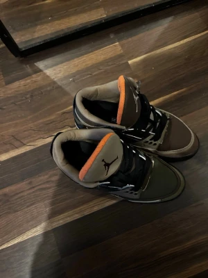 Nike Air Jordan 3 Camouflage - Unika Nike Air Jordan 3 sneakers med camouflagemönster i grönt, brunt och svart. Skorna har orange detaljer på plösen och sulan, svarta skosnören och en mix av textil och skinn. Klassisk Jordan-logga på plösen och robust sula med tydlig orange detalj undertill.