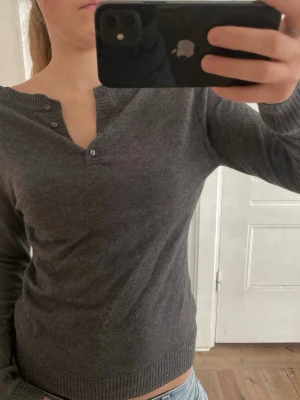 Mörkgrå stickad långärmad tröja - Säljer en mörkgrå stickad tröja med lång ärm och ribbade muddar. Tröjan har en v-ringad hals med knappdetaljer som ger en chill och enkel look. Perfekt att matcha med jeans eller kjol för en clean stil.