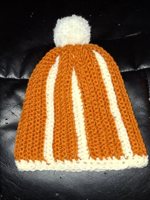 Randig orange och vit Virkad mössa - Säljer en handgjord stickad mössa i orange och vitt med breda ränder och en stor vit boll på toppen. Mössan har en klassisk form och är perfekt för kalla dagar. Passar dig som vill ha en färgglad och mysig accessoar. Strl S/M 