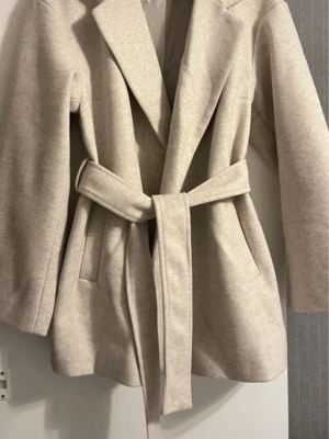 Beige kappa med bälte - Snygg beige kappa med breda slag och knytskärp i midjan. Jackan har raka fickor på sidorna och är långärmad. Perfekt för dig som gillar en stilren och enkel look. Materialet känns mjukt och har en ullkänsla.