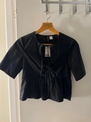 Svart blus med knyt från Divided - Säljer en svart blus från Divided med knyt framtill och rund hals. Blusen har korta ärmar och en lätt volang nedtill som ger en söt siluett. Den är helt oanvänd med till och med prislappen kvar 