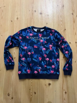 Blå sweatshirt med flamingos från Grand Frank - Säljer en blå sweatshirt från Grand Frank med färgglatt mönster av rosa flamingos och tropiska blad. Tröjan har rund hals, långa ärmar och ribbade muddar. Perfekt för dig som vill sticka ut med en unik och lekfull stil. Är storlek L men känns mer som M.
