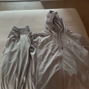 Grå tracksuit från Under Armour - Snygga grå mjukisbyxor från Under Armour med dragsko i midjan och dragkedjeförsedda fickor. Byxorna har en sportig look och är perfekta för chill eller träning. Materialet är mjukt och stretchigt för extra komfort.