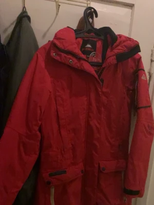 Röd parkasjacka från McKinley - Säljer en röd parkasjacka från McKinley med huva och praktiska fickor. Jackan har dragkedja och knappar framtill samt justerbara muddar vid ärmsluten. Perfekt för kalla dagar och outdoor-äventyr. Klassisk design med sportig känsla. 