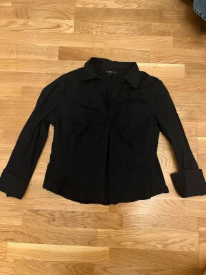 Svart skjorta från H&M, strl 46 - Klassisk svart skjorta från H&M i storlek 46. Skjortan har lång ärm, krage och knappar framtill. Tillverkad i ett mjukt bomullstyg som känns skönt mot huden. Perfekt till jeans eller kjol för en clean look.
