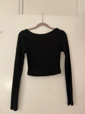 Svart ribbad croppad topp NA-KD med öppen rygg  - Säljer en svart långärmad topp från NA-KD i ribbat material. Toppen är croppad och har en öppen rygg med en snygg guldfärgad kedjedetalj. Perfekt att styla med höga jeans eller kjol för en trendig look.