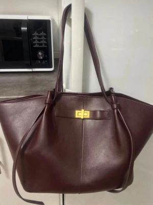  Vinröd axelväska i skinn med gulddetalj - Stilren brun axelväska i skinn med lång axelrem och snygg guldfärgad vridlåsdetalj framtill. Väskan har en rymlig form och minimalistisk design, perfekt för dig som vill ha plats för allt du behöver i vardagen.