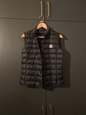 Svart dunväst från Moncler - Säljer en svart dunväst från Moncler med klassisk quiltad design och hög krage. Västen har tryckknappar framtill och Moncler-logga på bröstet. Perfekt att slänga över en hoodie eller tröja för en clean och sportig look. Använd fåtal gånger och ingen tidigare ägare! Nypris 9000kr