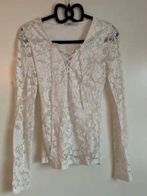 Vit spetsblus med snörning  - Säljer en vit långärmad blus från Gina Tricot i genombruten spets med blommönster. Toppen har v-ringning med snygg snörning framtill och är figurnära i modellen. Perfekt för dig som gillar romantiska detaljer och vill ha något extra till garderoben.