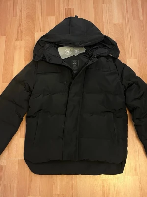 Svart pufferjacka från Canada Goose - Säljer en svart pufferjacka från Canada Goose med huva och diskret logga på ärmen. Jackan har dragkedja och tryckknappar framtill samt två stora fickor. Perfekt för kalla dagar och riktigt snygg till streetwear-stilen.