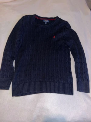 Mörkblå kabelstickad tröja från Ralph Lauren - Kabelstickad mörkblå tröja från Polo Ralph Lauren med klassisk röd logga broderad på bröstet. Tröjan har rund halsringning, ribbade muddar och är långärmad. Perfekt till lager på lager under höst och vinter. Varsamt använd, väldigt fint skick. Priset går att diskutera bara att skriva vid minsta fundering.