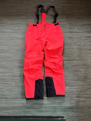 Orangea täckbyxor Whistler strl 42 - Säljer ett par orangea täckbyxor från Whistler med svarta förstärkningar vid bensluten och justerbara hängslen. Byxorna har dragkedja och knapp framtill, samt broderad logga och texten WX PRO 15000. Perfekta för vinterns alla äventyr! Några mindre fläckar i rumpan efter pulkaaktiviteter.