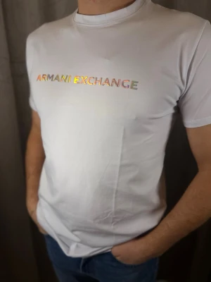 Vit t-shirt från Armani Exchange - Snygg vit t-shirt från Armani Exchange med holografiskt tryck på bröstet. Klassisk rund hals och korta ärmar. Tillverkad i mjuk bomull som känns skön mot huden. Perfekt för dig som gillar stilrena plagg med en twist.