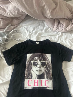 Svart t-shirt med CHIC-tryck - Svart t-shirt från river Island med stort tryck framtill som visar ett porträtt och texten CHIC i rosa. Klassisk rund hals och korta ärmar. Skön och mjuk bomullskänsla, perfekt till jeans eller kjol för en cool look. 