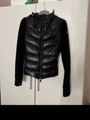 Svart pufferjacka från Moncler - Säljer en svart pufferjacka från Moncler med glansig front och huva. Ärmarna är i mjuk fleece och har en broderad Moncler-logga på vänster ärm. Jackan har dragkedja framtill och en schysst, sportig look. Perfekt för dig som vill ha både stil och komfort.