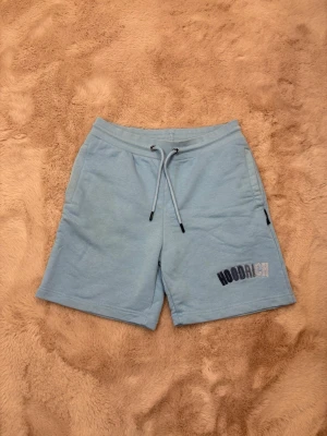 Hoodrich - Säljer ett par ljusblå shorts från Hoodrich i mjuk bomull. De har elastisk midja med snörning och fickor på sidorna. Hoodrich-loggan är broderad i svart och silver på ena benet. Perfekta för chill dagar eller när du vill ha en avslappnad look.