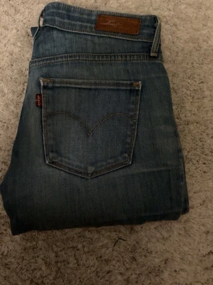 Levi's blå bootcut jeans Demi Curve - Säljer ett par klassiska blå Levi's Demi Curve jeans med bootcut. Jeansen har fickor och en mörkblå tvätt. Tillverkade i slitstarkt jeanstyg med den ikoniska röda Levi's-lappen på bakfickan och läderpatch i midjan.