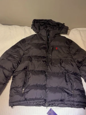 Svart pufferjacka från Polo Ralph Lauren - Säljer en svart pufferjacka från Polo Ralph Lauren med huva och dragkedja. Jackan har det klassiska röda Polo-logot på bröstet, flera fickor med dragkedja och en schysst oversized passform. Perfekt för kalla dagar och riktigt snygg till streetwear-stilen. Helt ny!!