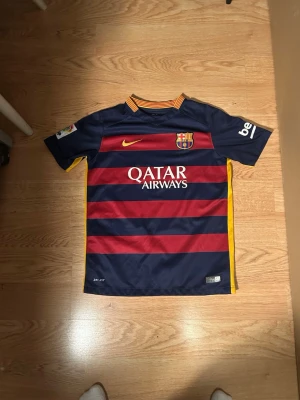 Barça matchtröja Nike Dri-FIT randig - Säljer en FC Barcelona matchtröja från Nike med Dri-FIT-teknologi. Tröjan har breda marinblå och röda ränder, gula detaljer på sidor och krage samt klubbmärke och Qatar Airways-tryck på bröstet. Perfekt för dig som älskar fotboll och Barça!
